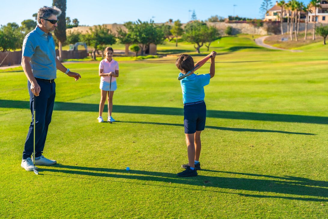 784304 Black Iron Golf | Diario-ABC.Com Boy in blue shirt swings a golf club while a man watches on a sunny course; a girl stands with a club in the background.