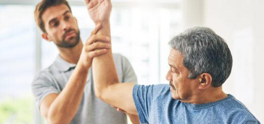 Physical therapist guiding a senior man through an arm-raising exercise in a bright room