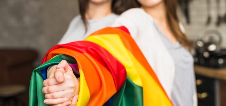 Two women smiling and extending a rainbow pride flag toward the camera, symbolizing LGBTQ+ solidarity.