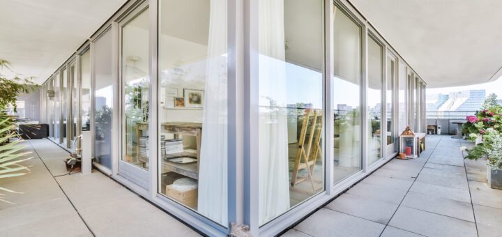 Glass-walled studio on a balcony with white curtains, visible art supplies, and easels inside; outdoor plaza and plants outside.