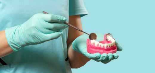 Dental professional in teal gloves holds a pink dental model while using a mirror tool to examine teeth prompt. (Informative/illustrative)