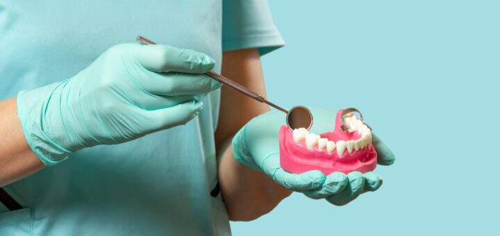 Dental professional in teal gloves holds a pink dental model while using a mirror tool to examine teeth prompt. (Informative/illustrative)