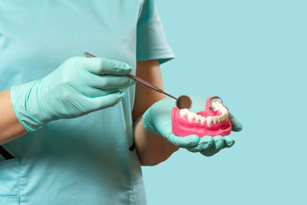 Dental professional in teal gloves holds a pink dental model while using a mirror tool to examine teeth prompt. (Informative/illustrative)