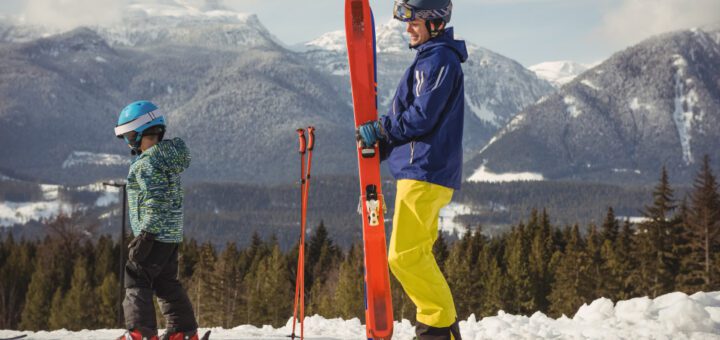 Adult in a blue jacket and bright yellow pants carries a red snowboard while a child in a teal helmet adjusts skis on a snowy mountain slope, with pine trees in the background.
