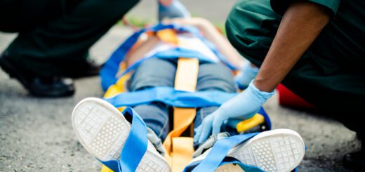 Emergency responders strap a person onto a yellow stretcher using blue straps, with gloved hands visible on a paved outdoor surface.