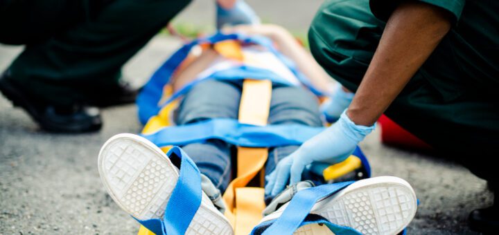 Emergency responders strap a person onto a yellow stretcher using blue straps, with gloved hands visible on a paved outdoor surface.