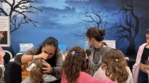 Children sit at a table getting Halloween makeup from two adults in a mall, with a blue Halloween backdrop reading 'HALLOWEEN GRAN PLAZA' behind them