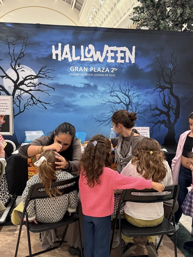 Halloween Gran Plaza 2 - IP | Diario-ABC.Com Children sit at a table getting Halloween makeup from two adults in a mall, with a blue Halloween backdrop reading 'HALLOWEEN GRAN PLAZA' behind them